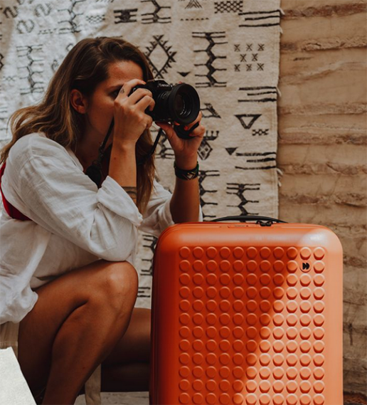 orange dot drops suitcase with woman looking through a camera